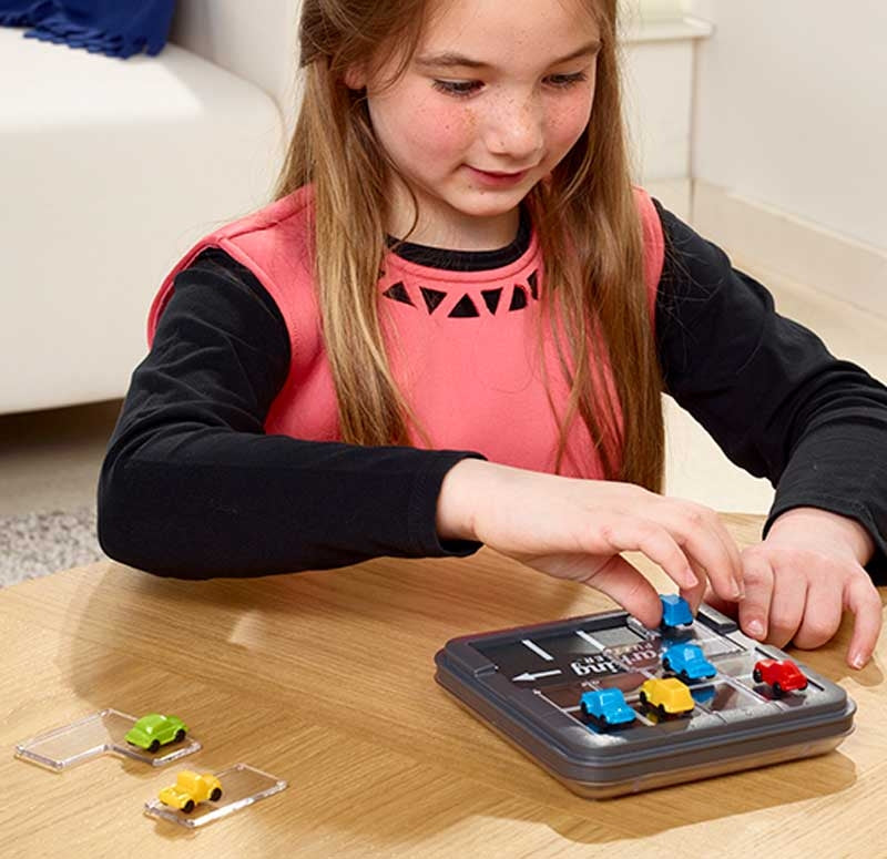 "A girl engaged in solving a challenge on the SmartGames Parking Puzzler, carefully placing colourful car pieces on the compact game board to complete the logic puzzle.