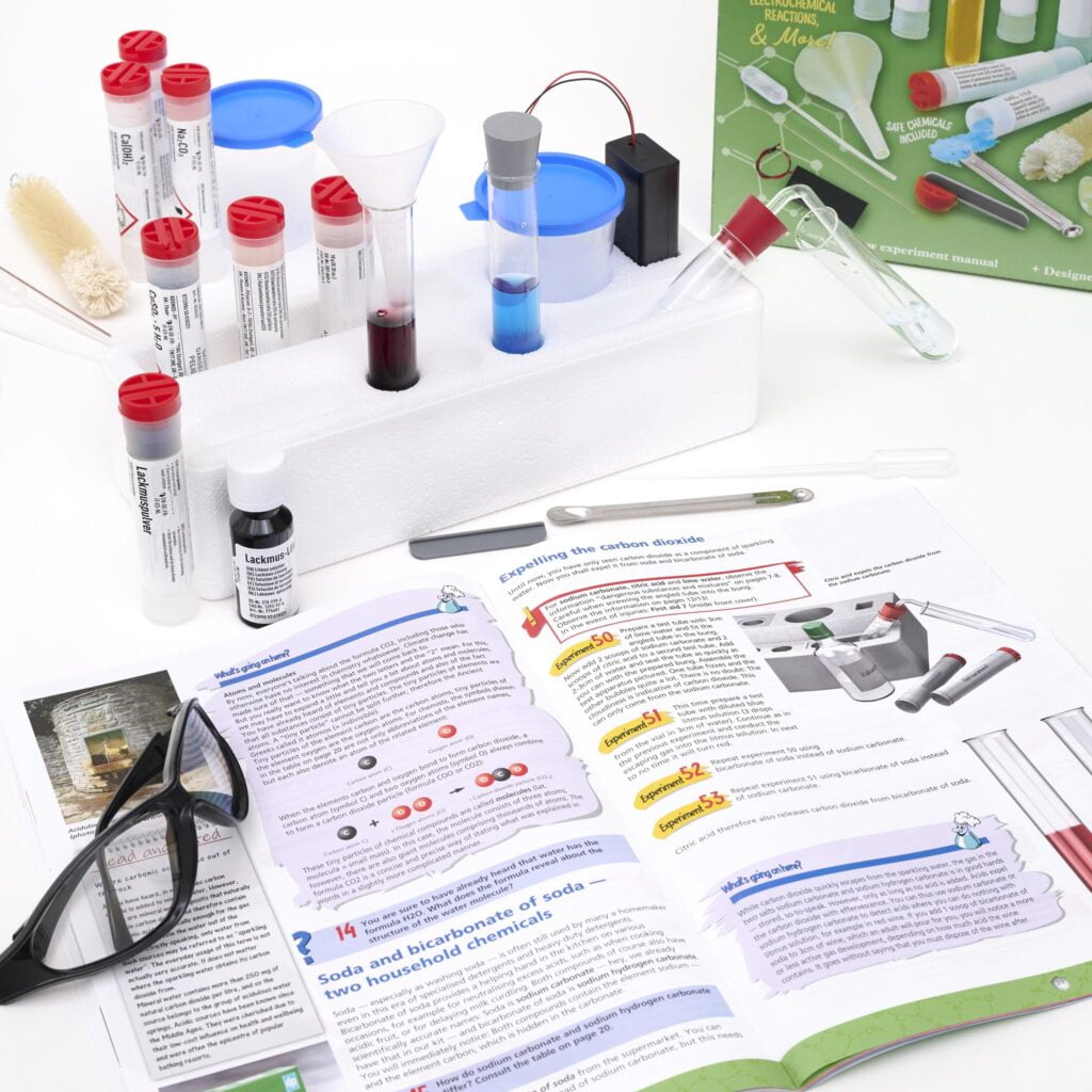 Thames & Kosmos Chemistry C1000 Set instruction book open with scattered chemistry items. Hands-on STEM learning with step-by-step experiments for young scientists.