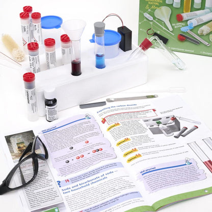 Thames & Kosmos Chemistry C1000 Set instruction book open with scattered chemistry items. Hands-on STEM learning with step-by-step experiments for young scientists.
