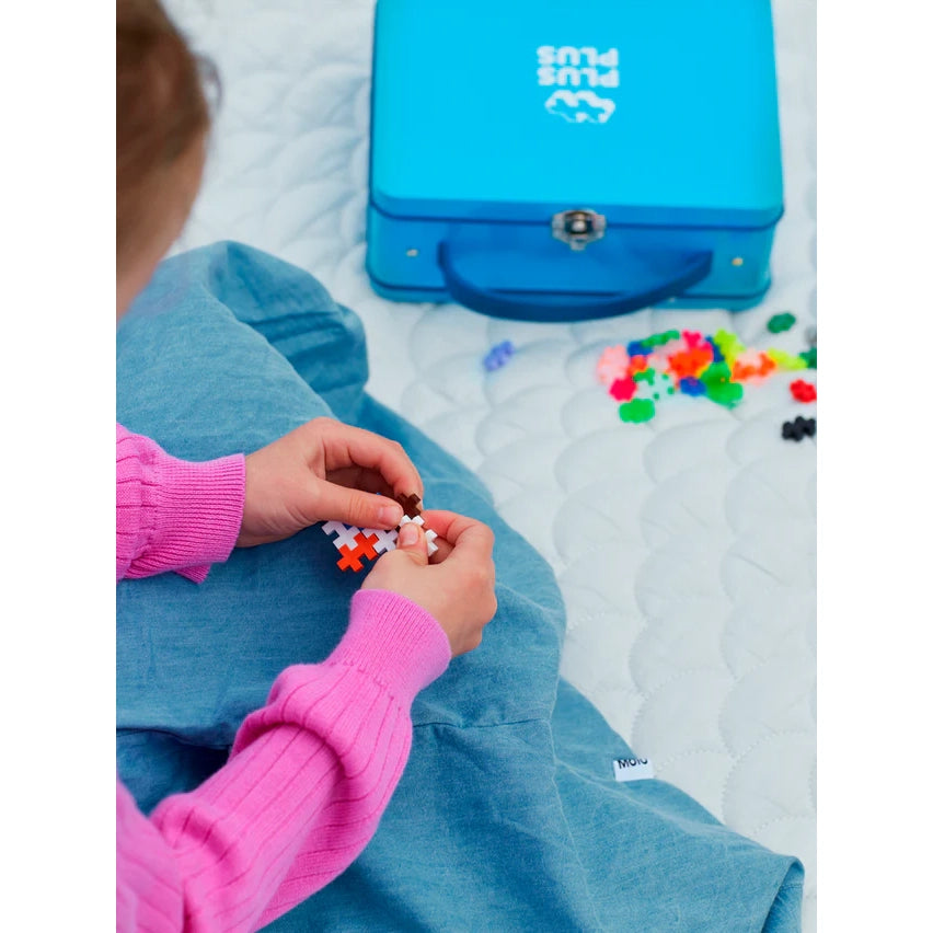A child building with colourful Plus-Plus pieces, with the blue metal suitcase nearby. The sturdy tin keeps 600 interlocking pieces organized for creative play