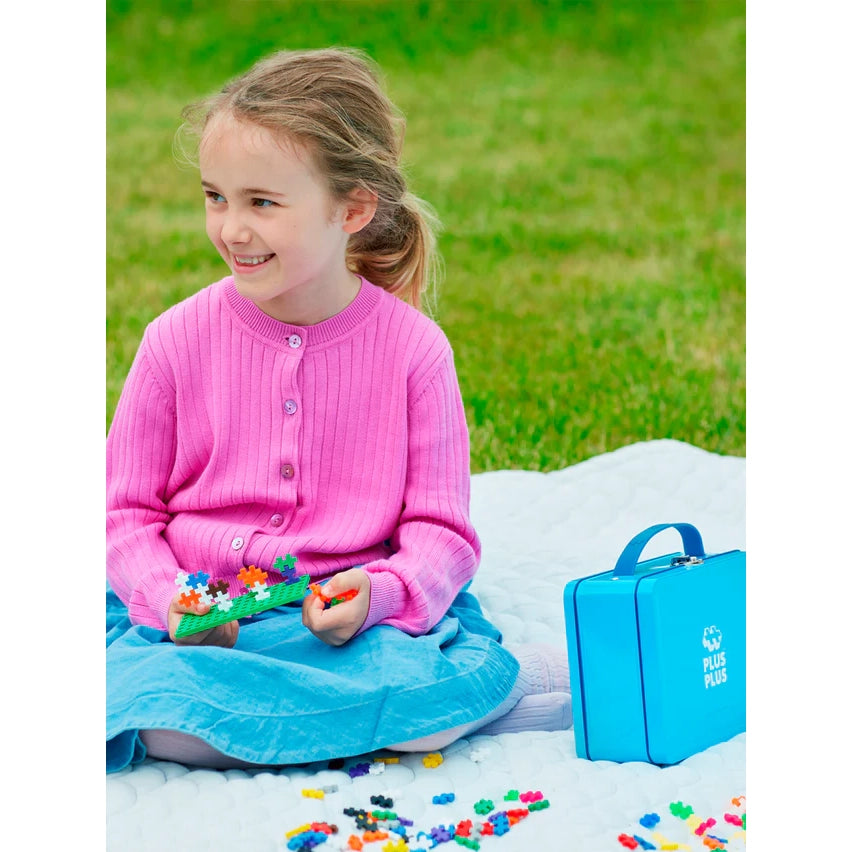 A child building with Plus-Plus pieces outside, with the blue metal suitcase nearby. The sturdy tin keeps 600 interlocking pieces organized for creative play on the go