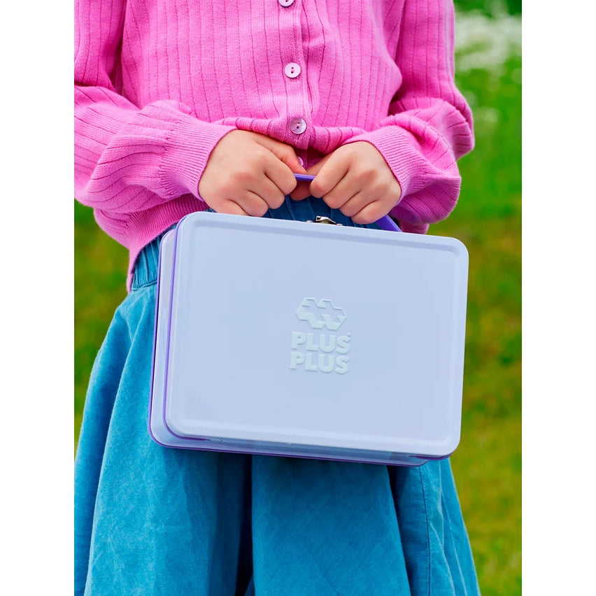 A smiling girl with her pastel Plus-Plus metal suitcase, surrounded by colourful built models made from interlocking pieces, ready for creative play.