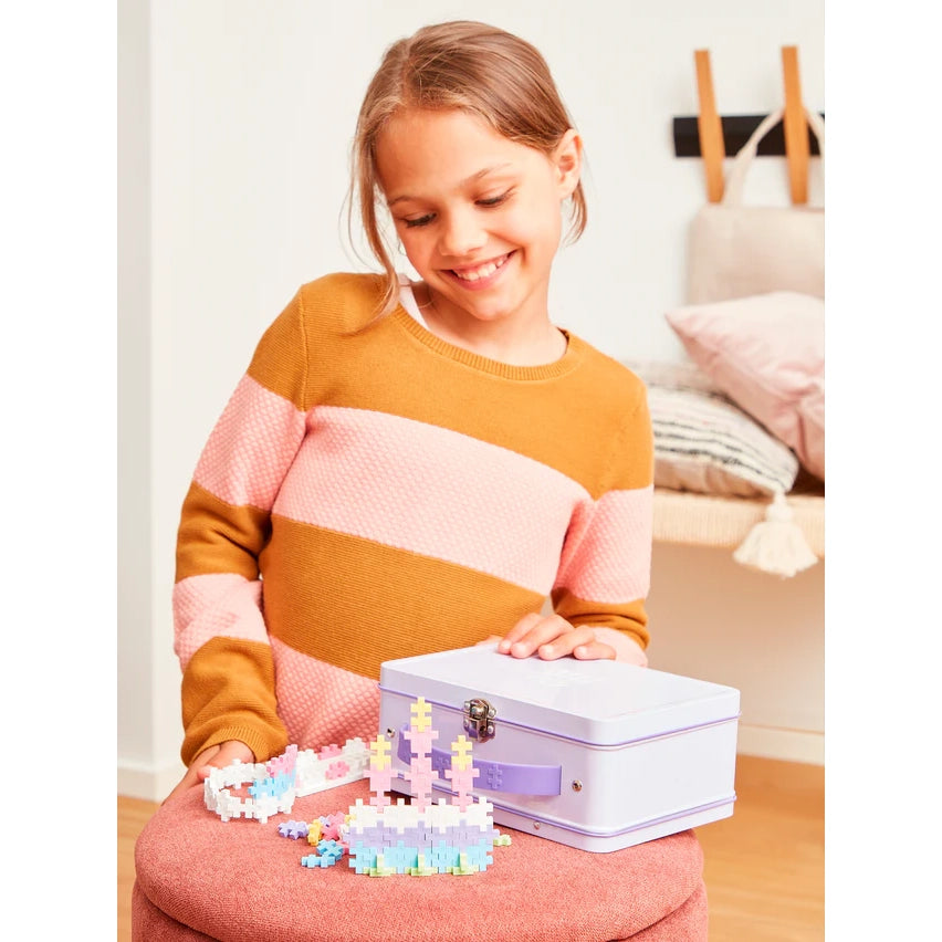 A smiling girl with her pastel Plus-Plus metal suitcase, surrounded by colourful built models made from interlocking pieces, ready for creative play.