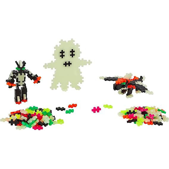 Three built models made from glow-in-the-dark Plus-Plus pieces, including a glowing ghost, with additional loose interlocking pieces scattered nearby